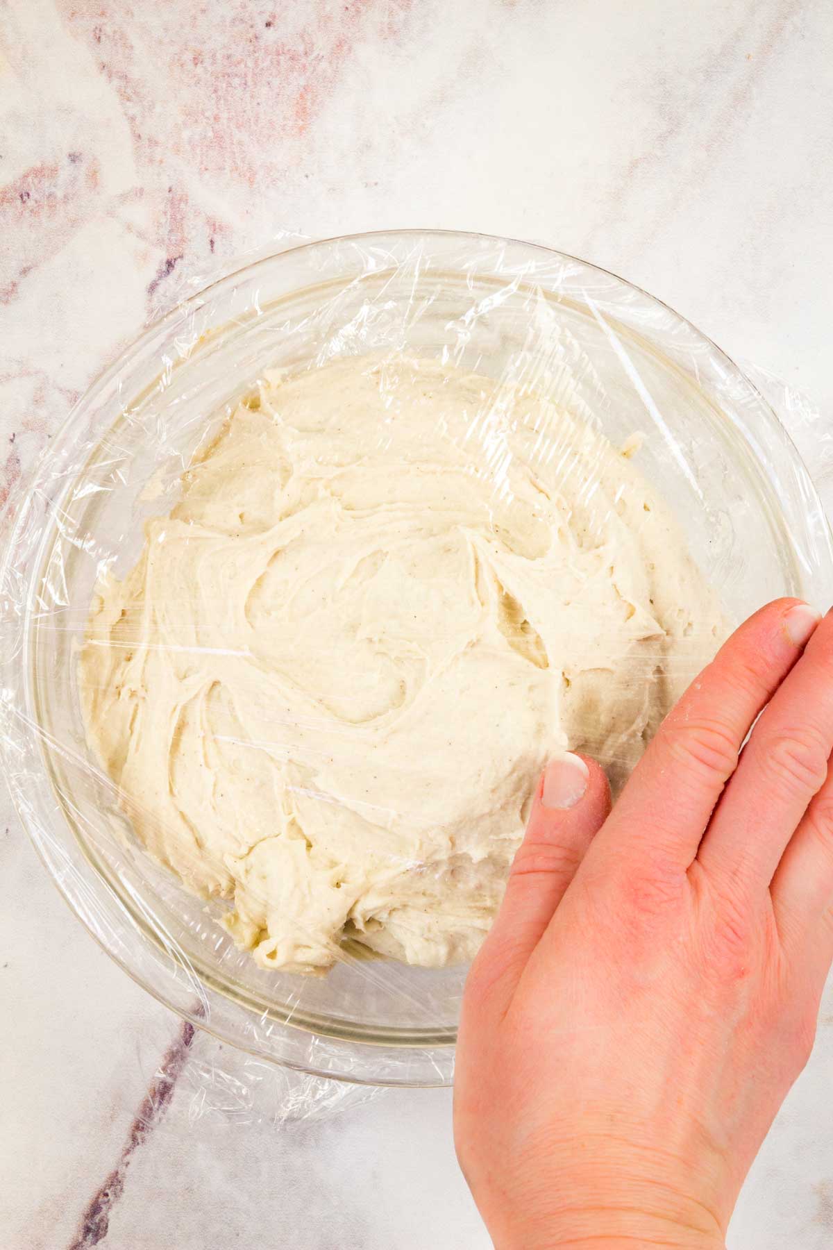 A hand covers a bowl of English muffin dough with plastic wrap.