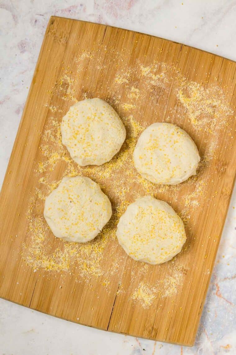 Shaped English muffin dough on a wooden cutting board, dusted with cornmeal.