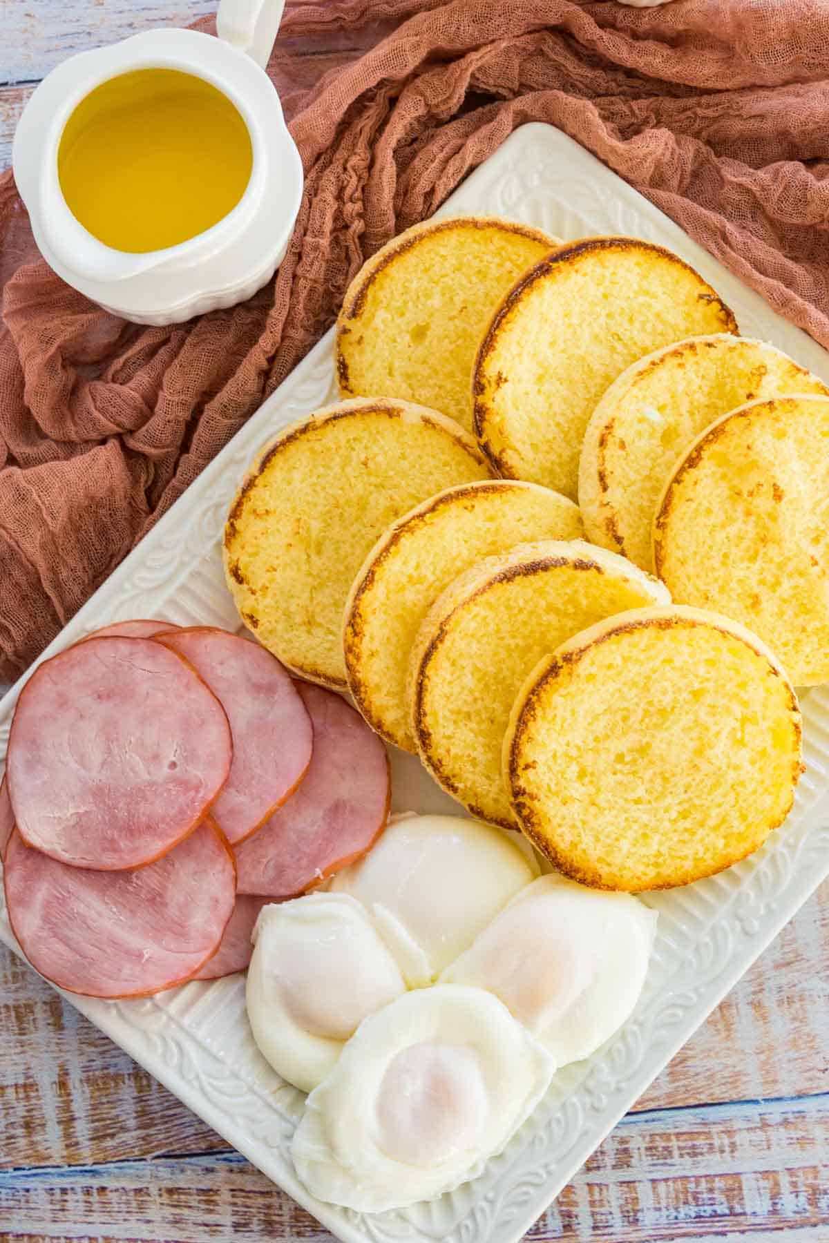 A serving tray filled with poached eggs, Canadian bacon, and English muffin halves, next to a boat of hollandaise sauce.