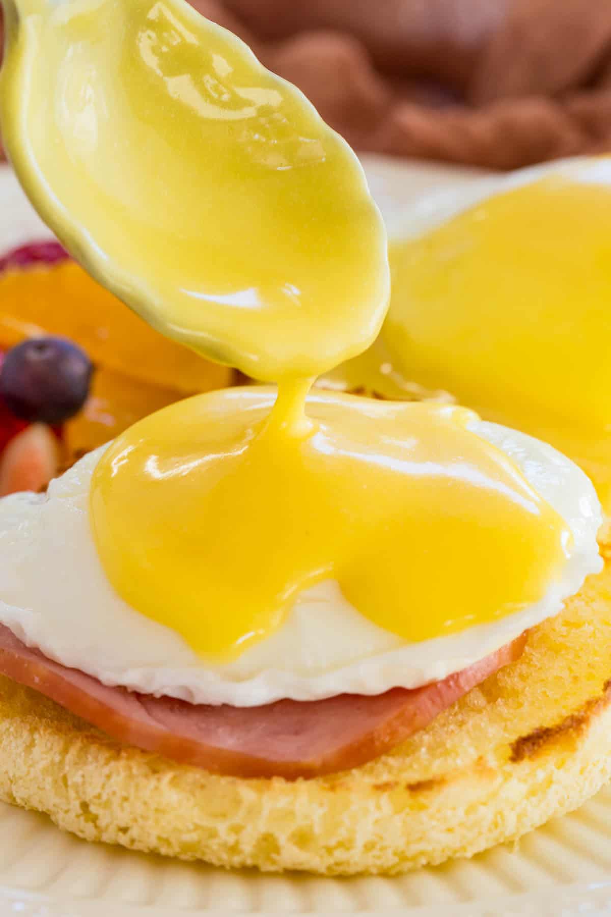A spoonful of hollandaise sauce is dropped onto the poached egg of an Eggs Benedict stack.