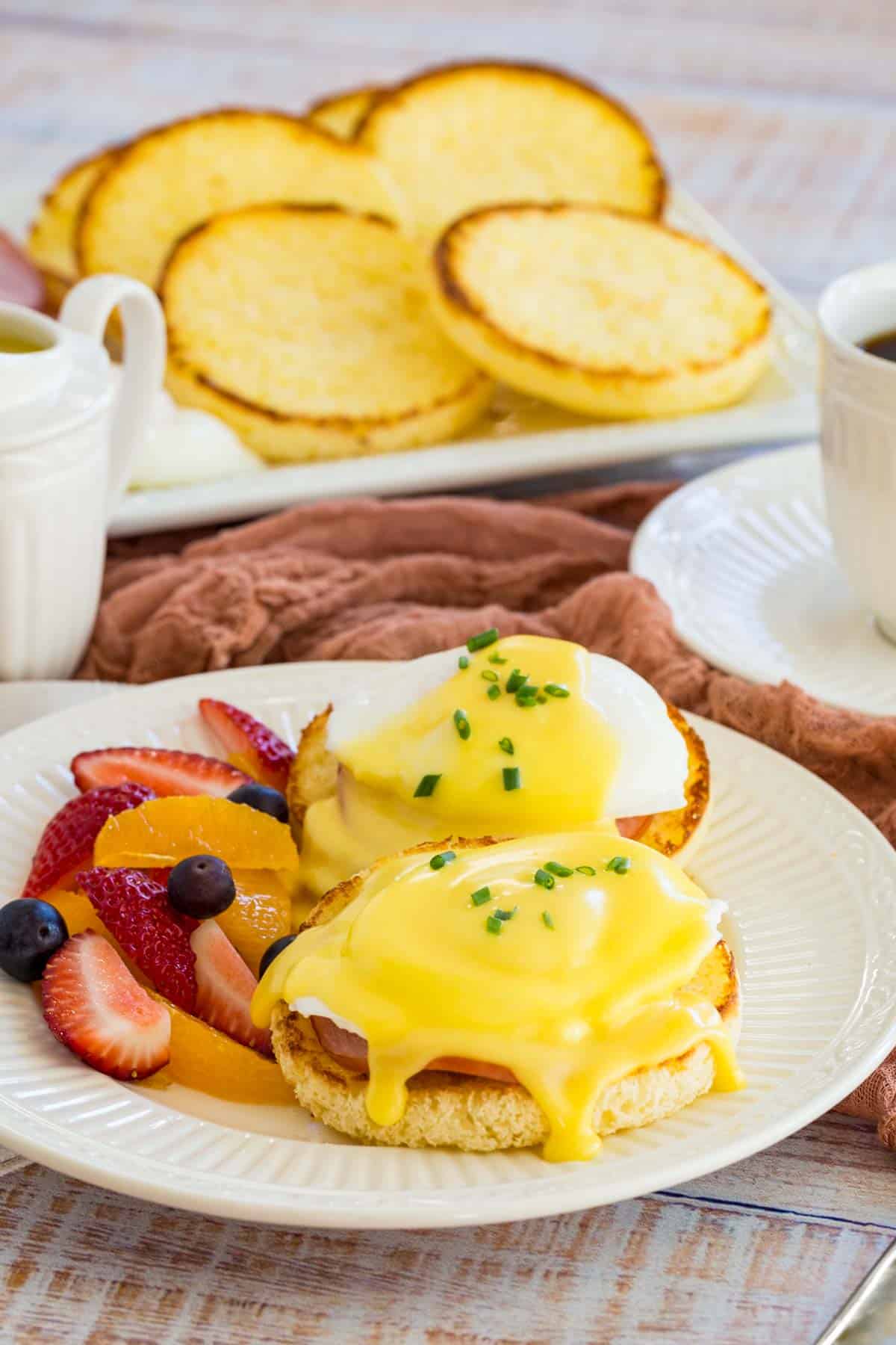 A brunch spread featuring a plate with Eggs Benedict and fresh fruit, with a tray filled with English muffins in the background, next to a cup of coffee.