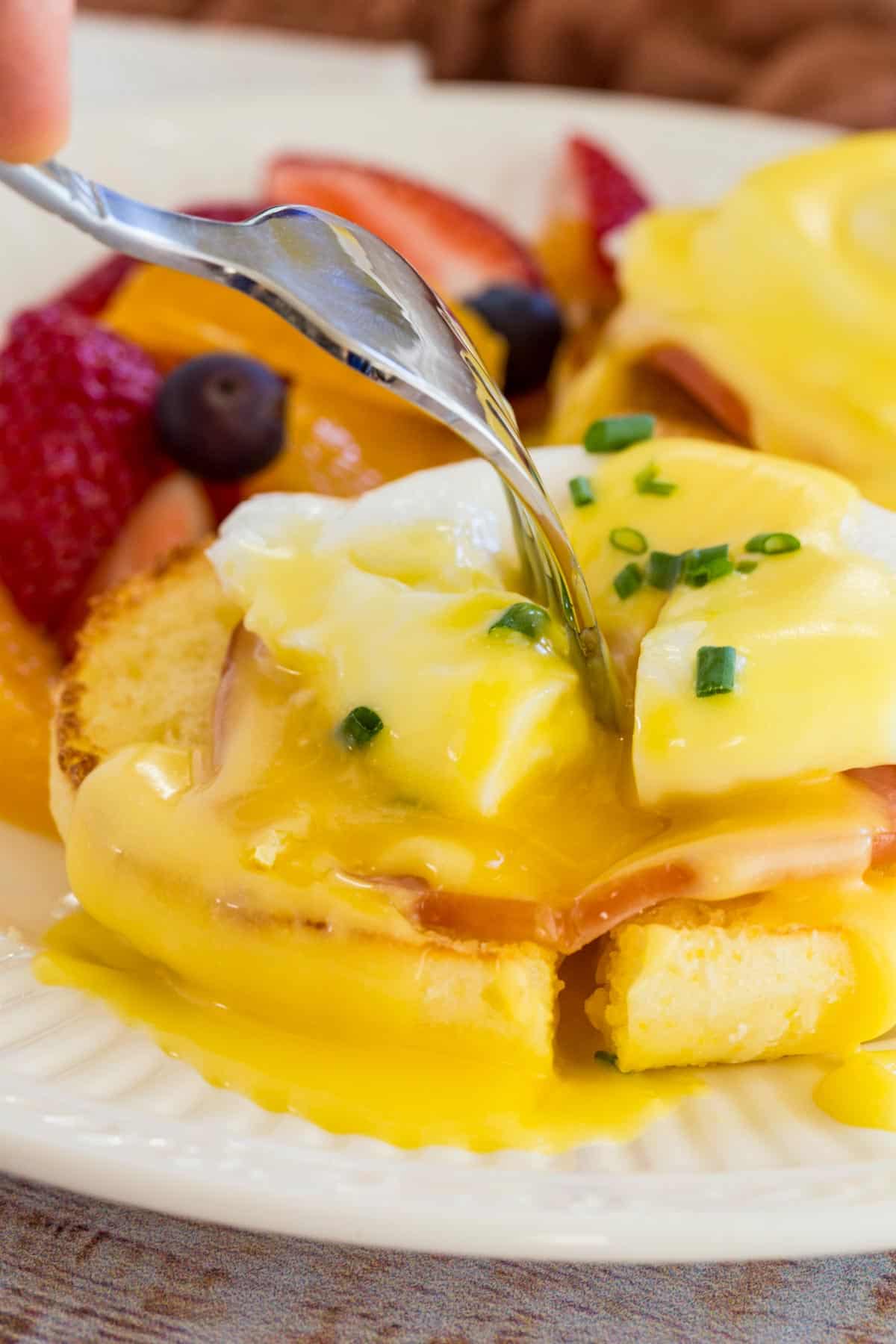 A fork is used to split open the poached egg on top of an Eggs Benedict stack.