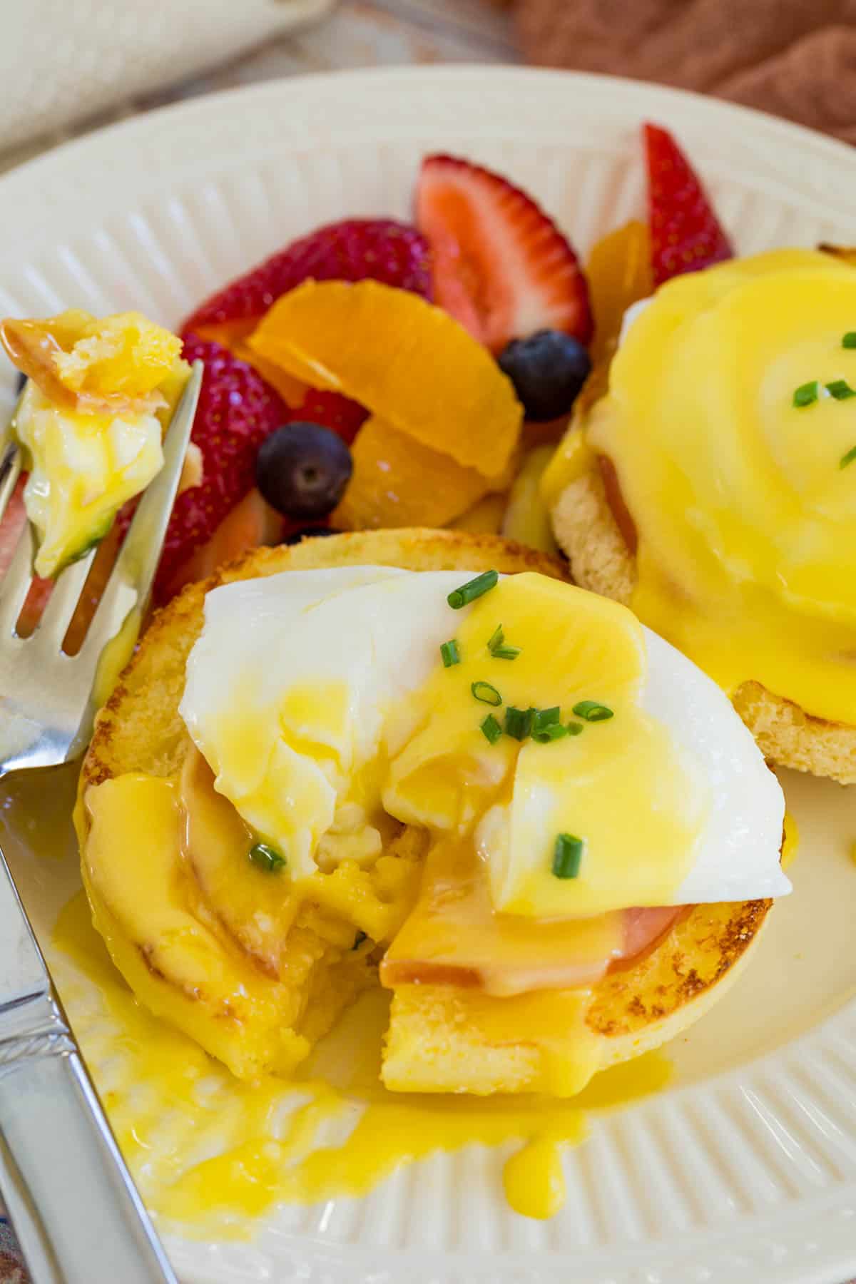 An Eggs Benedict stack that's been cut in half with a fork, with the egg yolk running, surrounded by fresh fruit.