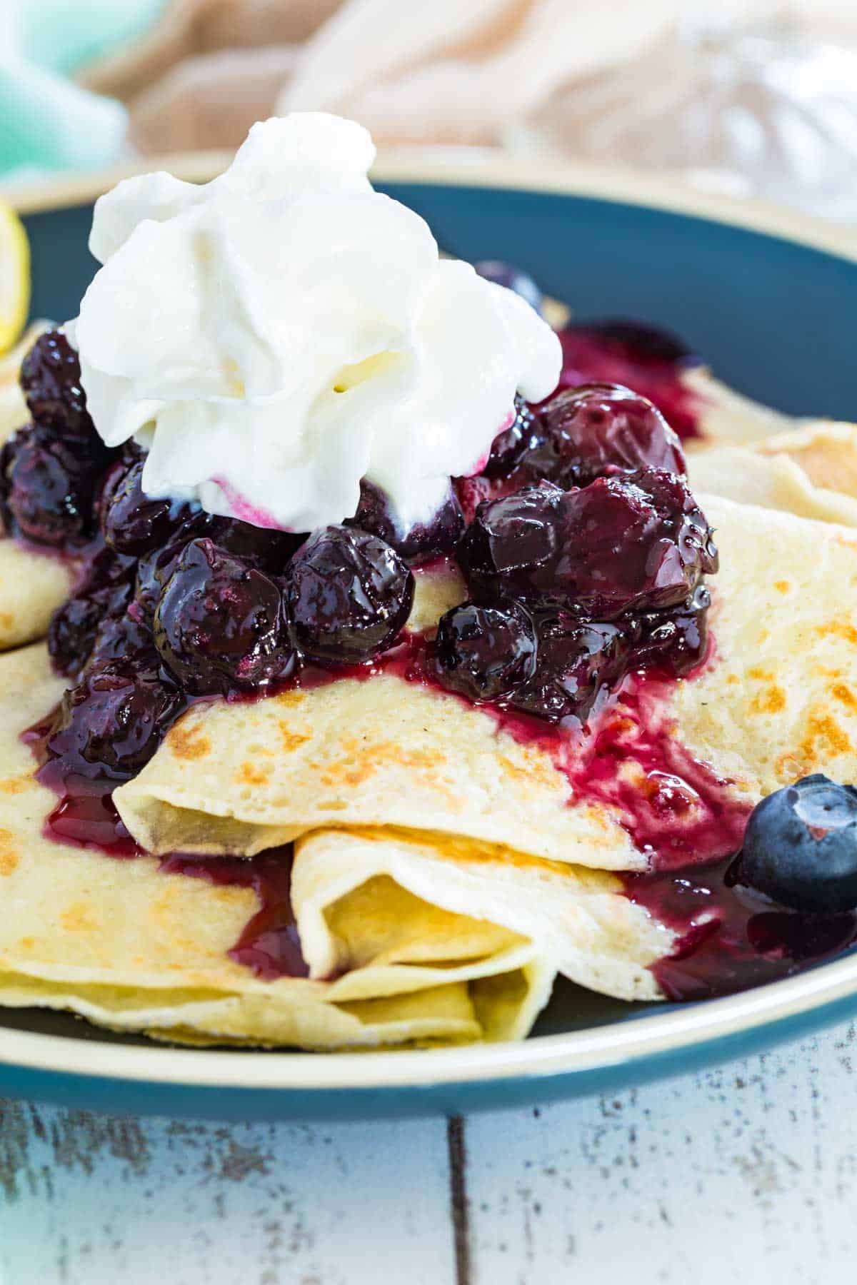 A close-up image of homemade crepes topped with blueberry sauce and whipped cream