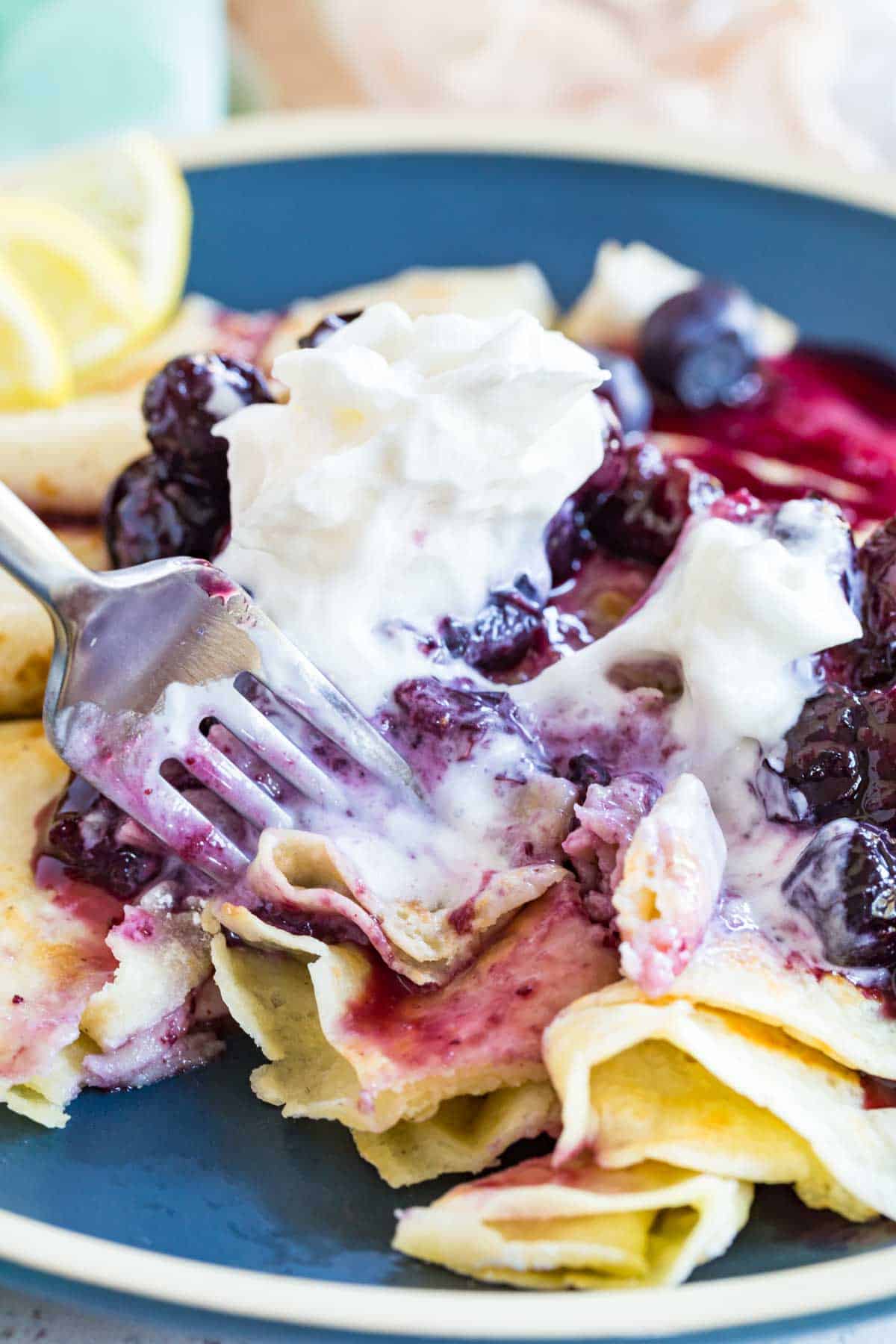 A serving of gluten-free blueberry crepes topped with melty whipped cream being poked with a metal fork