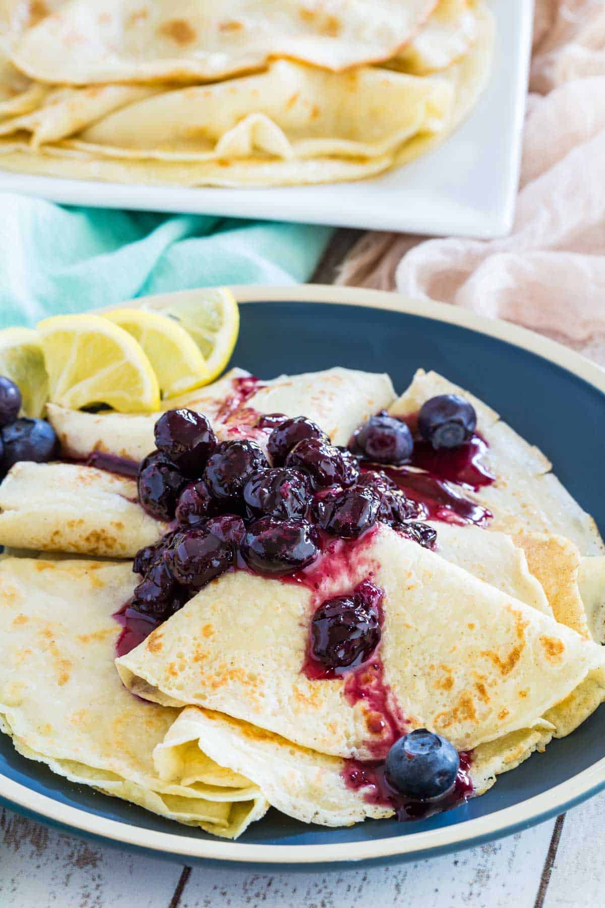 A pile of blueberry crepes on a plate with more crepes on a serving platter in the background