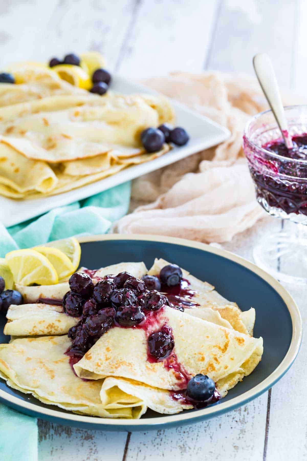 Gluten-free blueberry crepes on a plate with blueberry sauce and more crepes behind them
