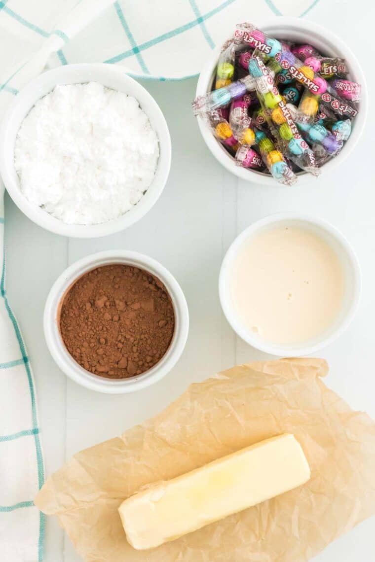 The ingredients to make the chocolate frosting plus a bowl of Sixlets for decorating on a countertop.