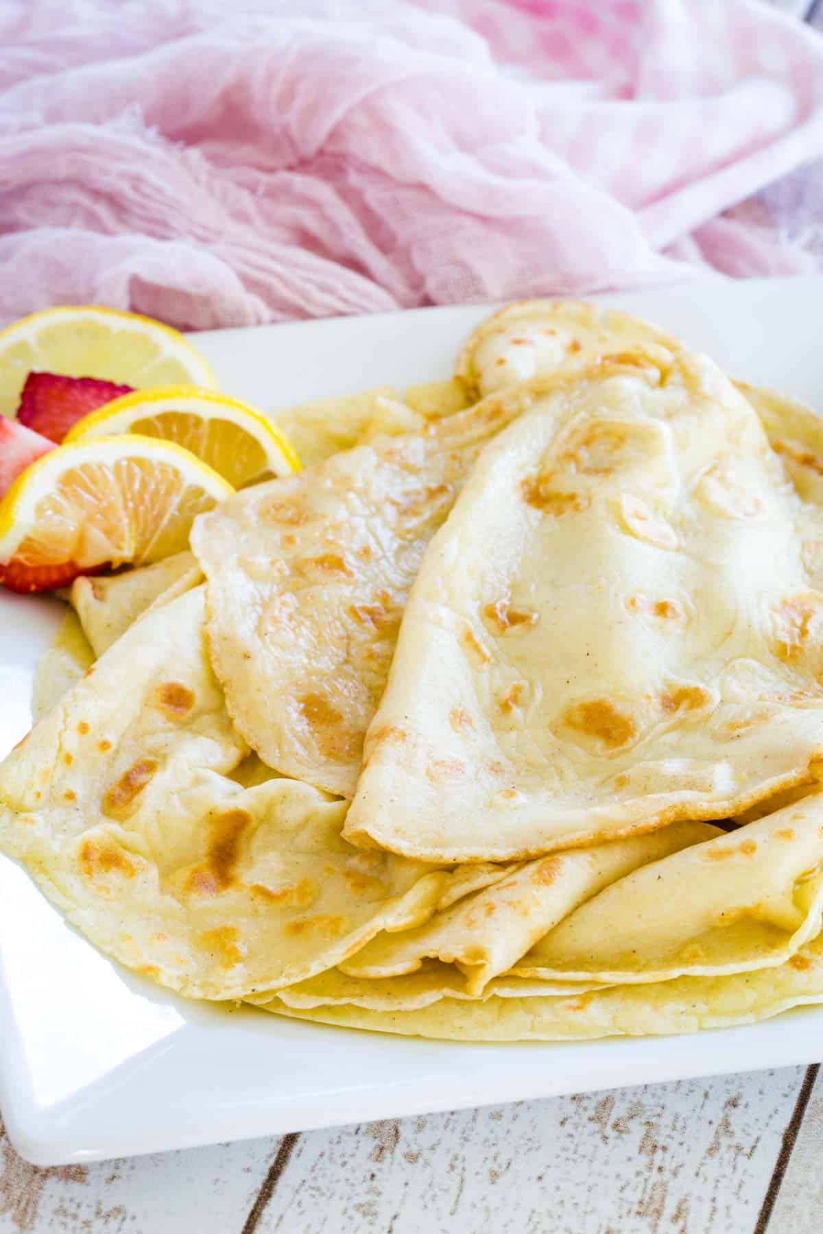 Crepes piled up on a white plate with some strawberry and lemon slices.