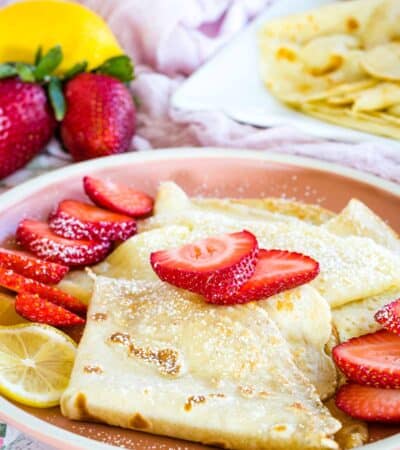 Gluten free crepes with strawberry and lemon slices and a dusting of powdered sugar on a pink plate.