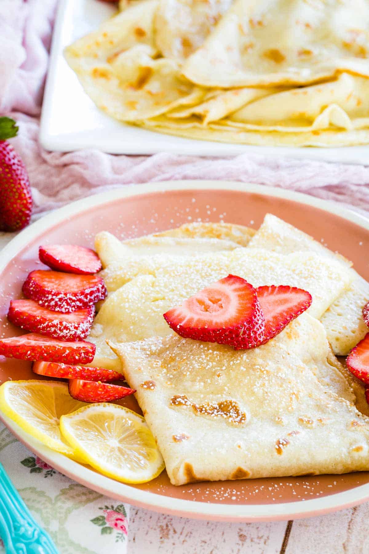Gluten free crepes on a pink plate topped with strawberries, with more crepes on a serving tray in the background.