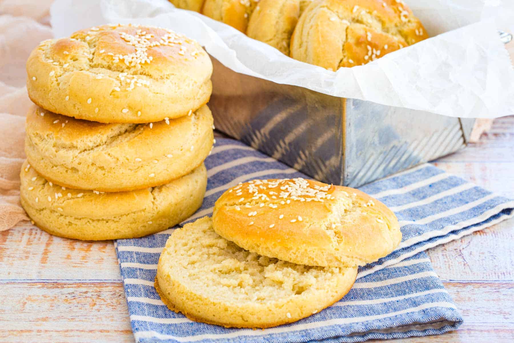 One slice hamburger bun on a striped napkin with three stacked and more in a pan.