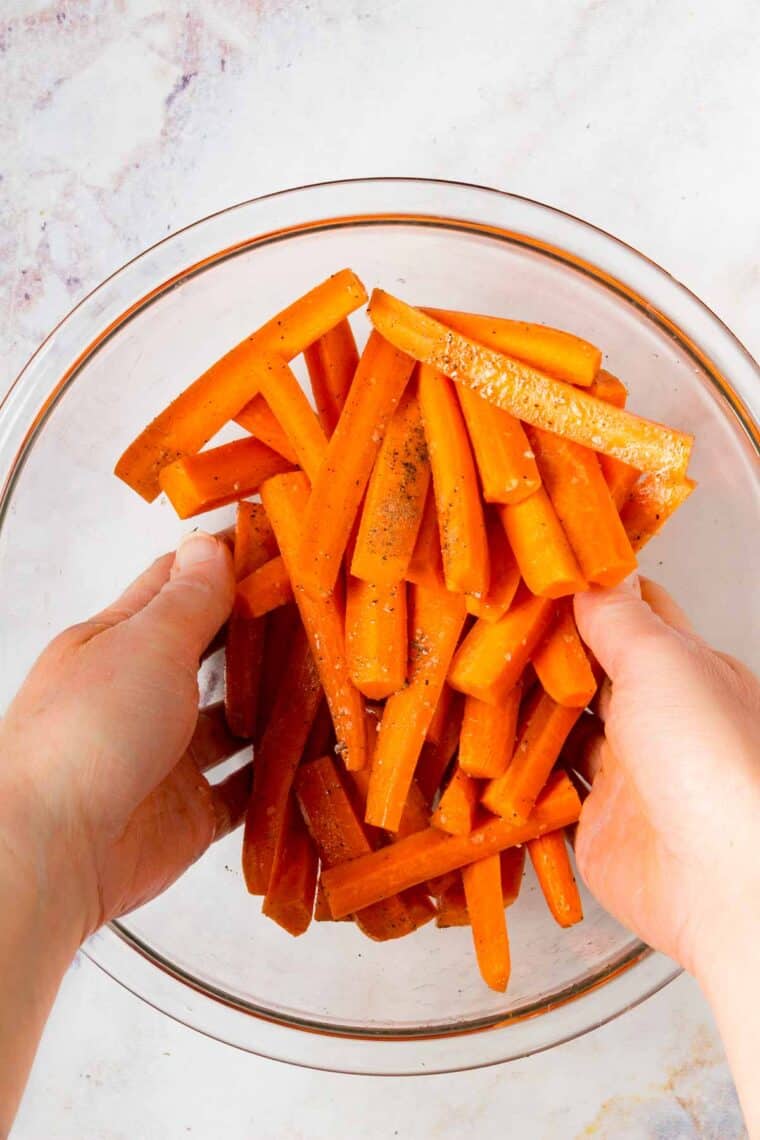Hands toss carrots in a bowl with oil and seasonings.