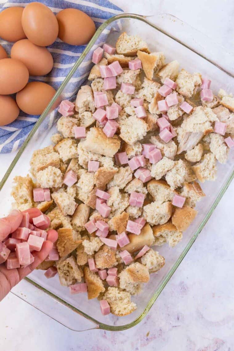 Chopped ham is sprinkled over chunks of gluten free baguette in a clear baking dish.