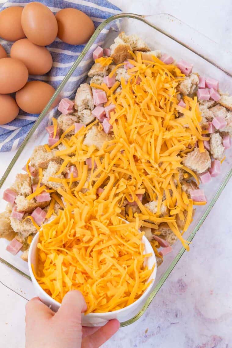 A layer of shredded orange cheddar is sprinkled over bread chunks and ham in a baking dish.