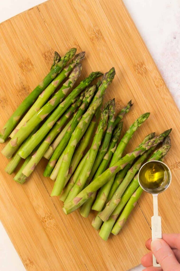 A tablespoon of olive oil is held over a pile of asparagus spears on a wooden cutting board.