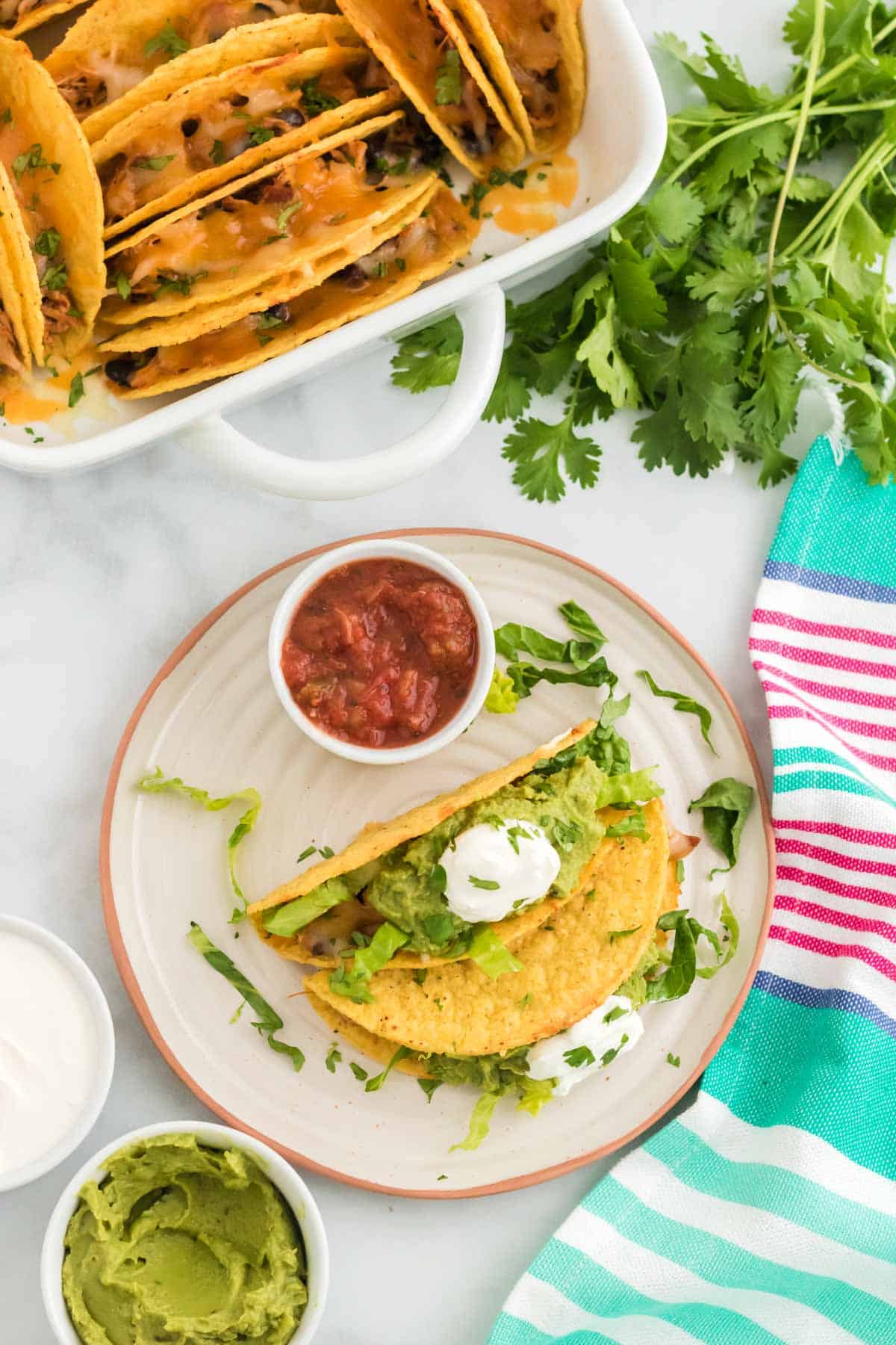 A plate with two baked chicken tacos and a bowl of salsa, next to toppings and a baking tray of tacos.