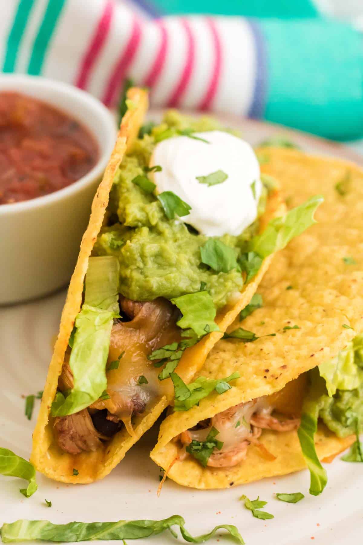 Two baked chicken tacos filled with avocado and sour cream on a plate, next to a bowl of salsa.