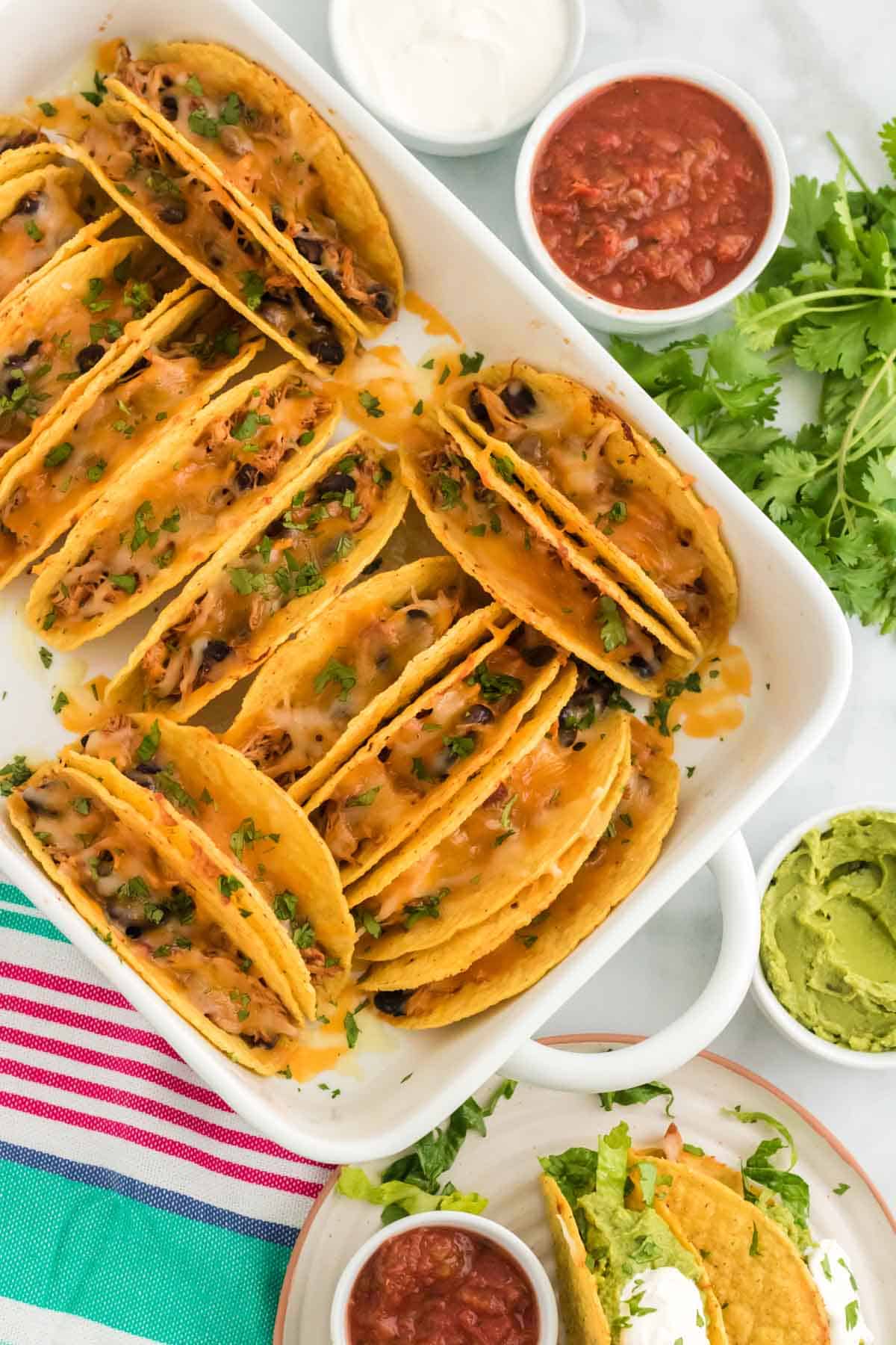 Top view of baked chicken tacos in a white baking dish surrounded by toppings and a plate with tacos..