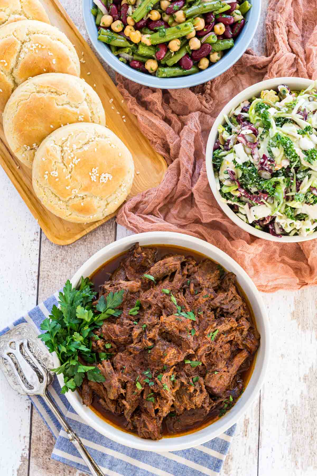 Barbecue beef in a white bowl next to bowls of coleslaw, three bean salad, and a tray of gluten-free buns.