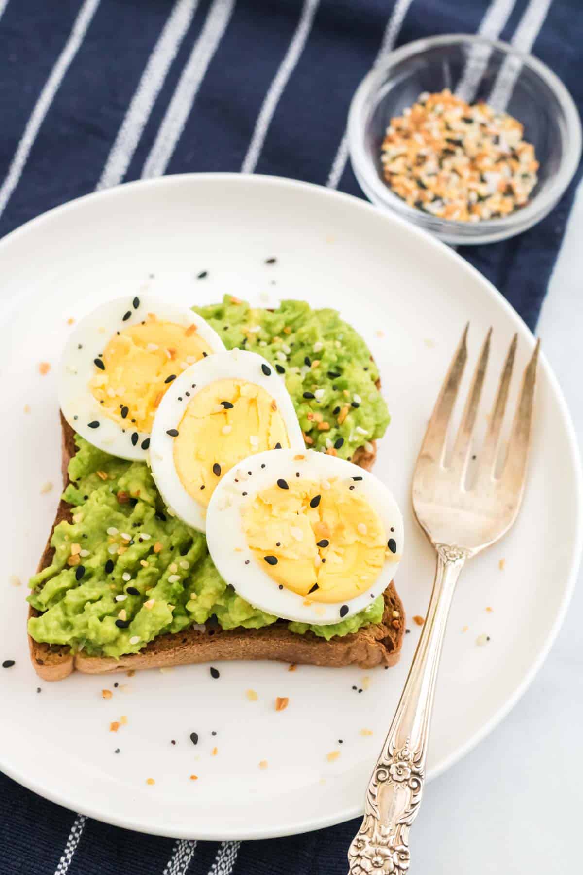 Avocado toast topped with sliced eggs and seasoning on a plate with a fork.