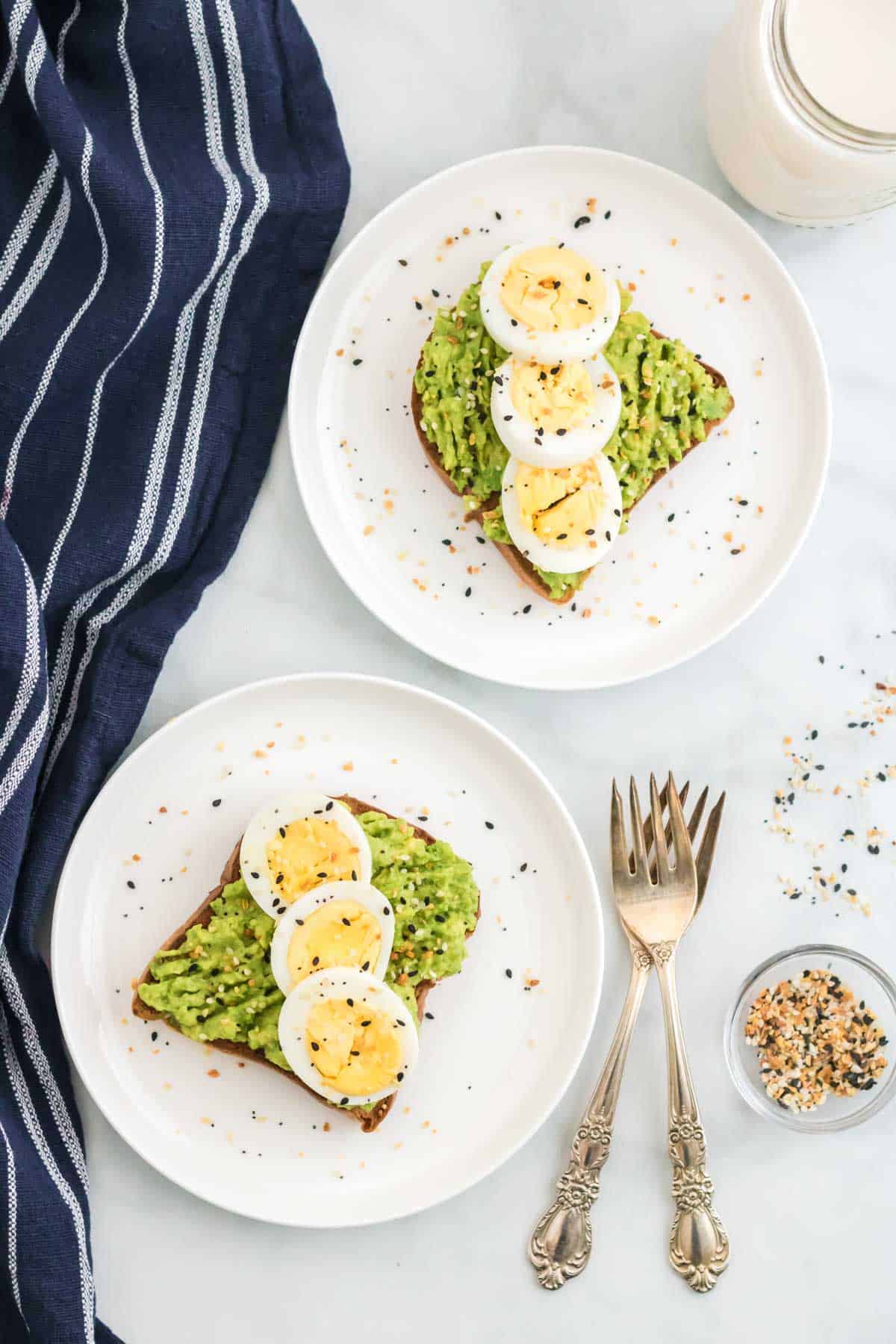 Avocado toasts topped with sliced hard boiled eggs and Everything Bagel seasoning, on two white plates next to two forks.