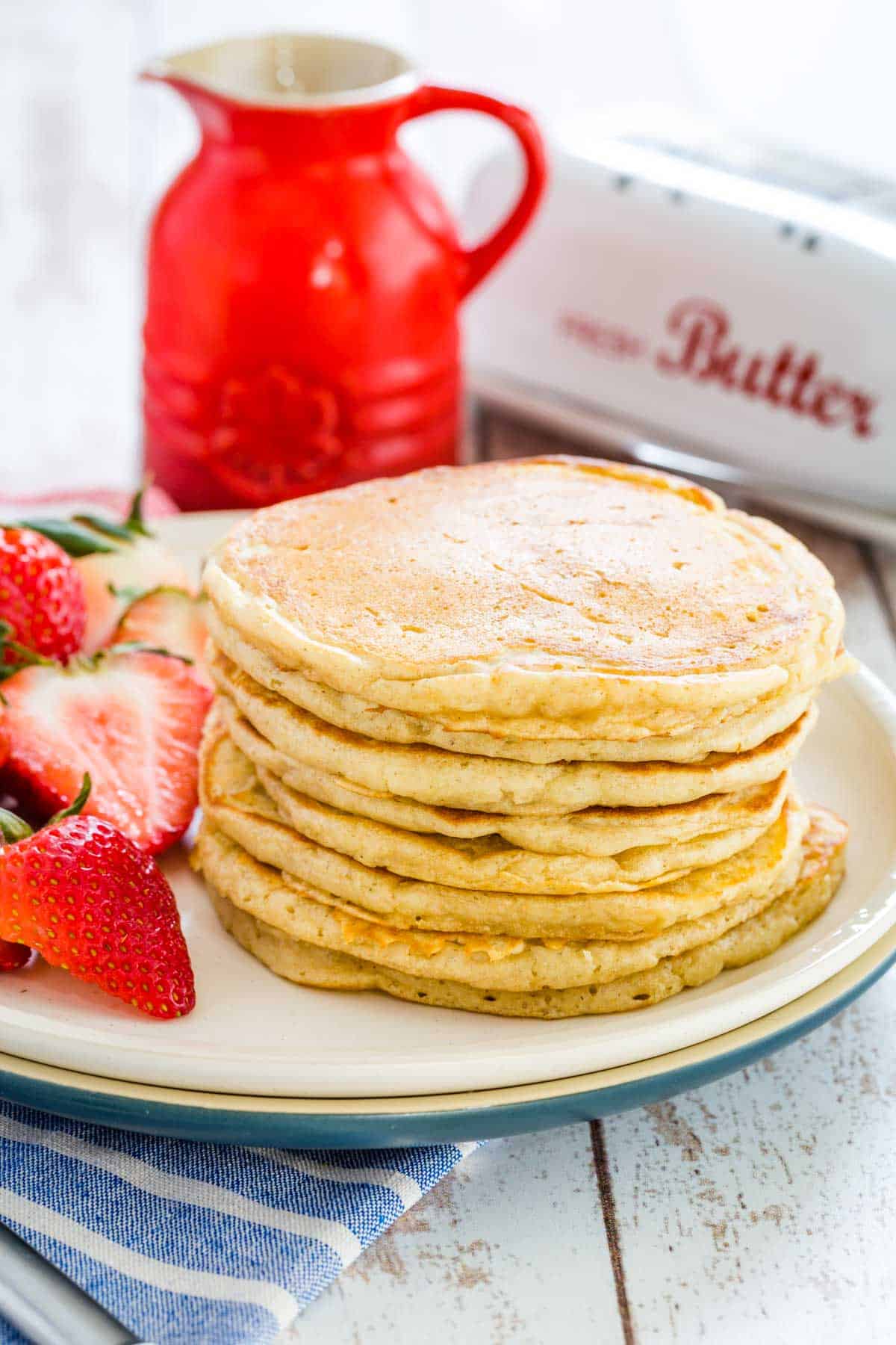 A stack of gluten free buttermilk pancakes on a plate next to sliced strawberries and a jug of maple syrup.