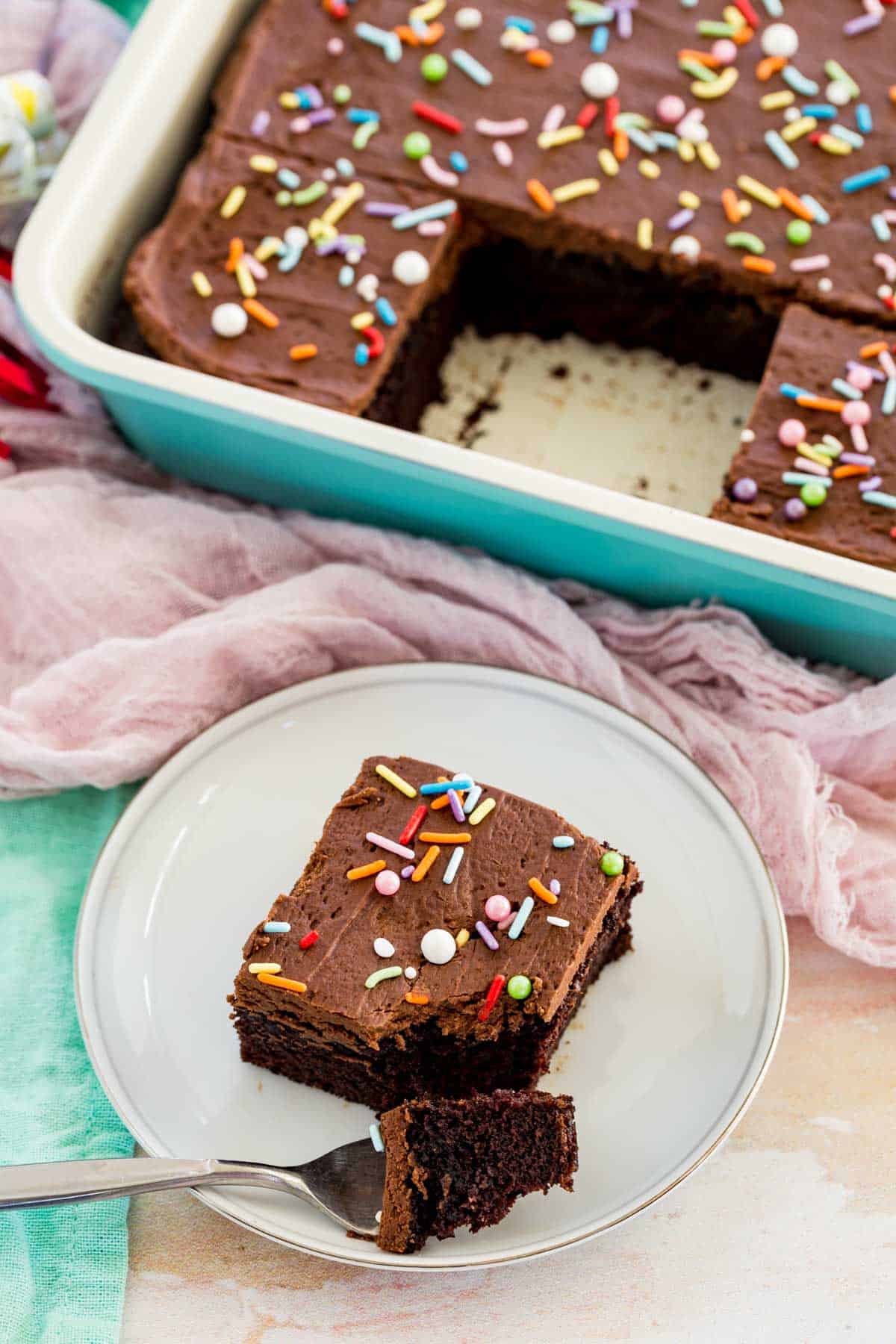 A square slice of chocolate cake with sprinkles on a plate, next to a baking dish filled with cake.