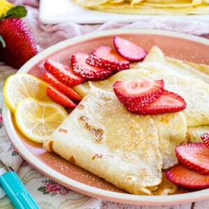 Gluten free crepes with strawberry and lemon slices and a dusting of powdered sugar on a pink plate.