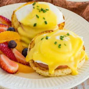 Two eggs benedict garnished with chives on a plate with fresh fruit.