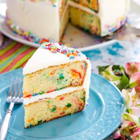 A slice of gluten free funfetti cake on a blue plate next to a fork, with the whole cake in the background.