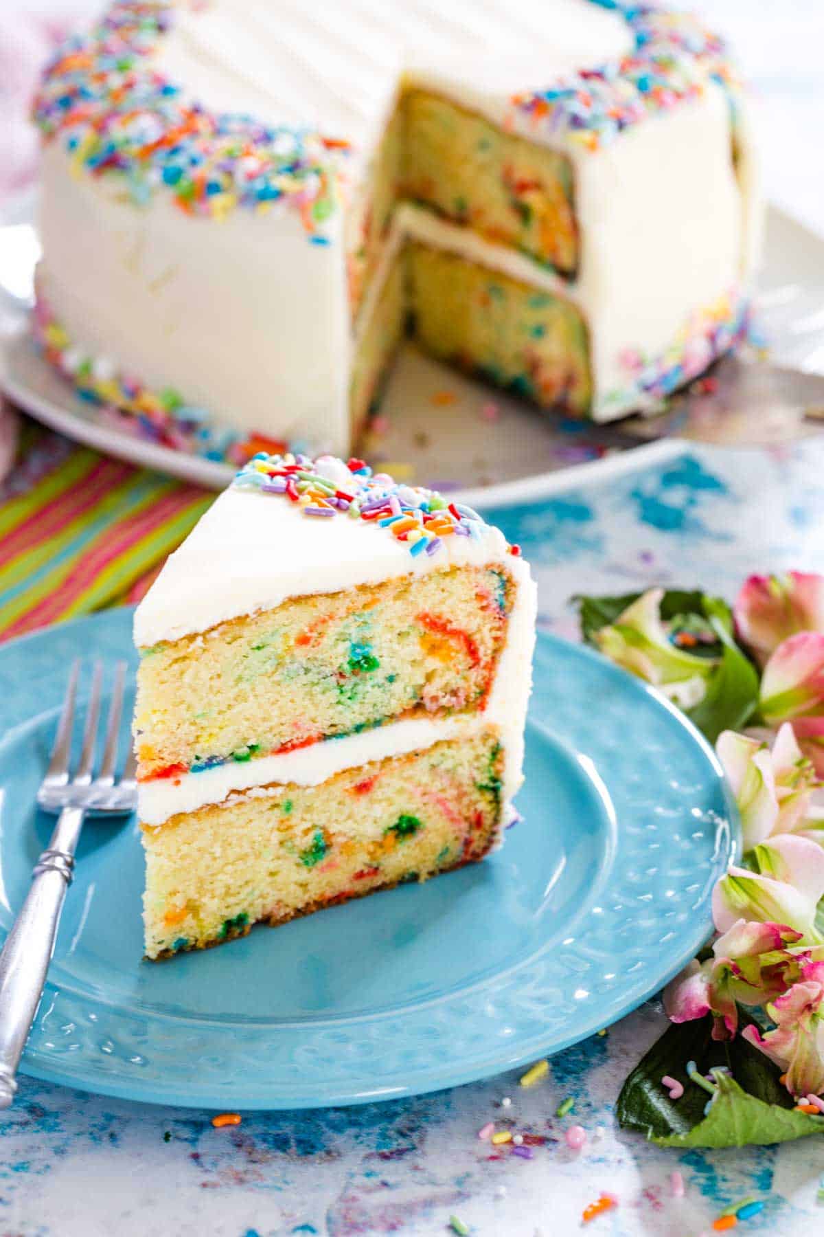 A slice of gluten free funfetti cake on a blue plate next to a fork, with the whole cake in the background.
