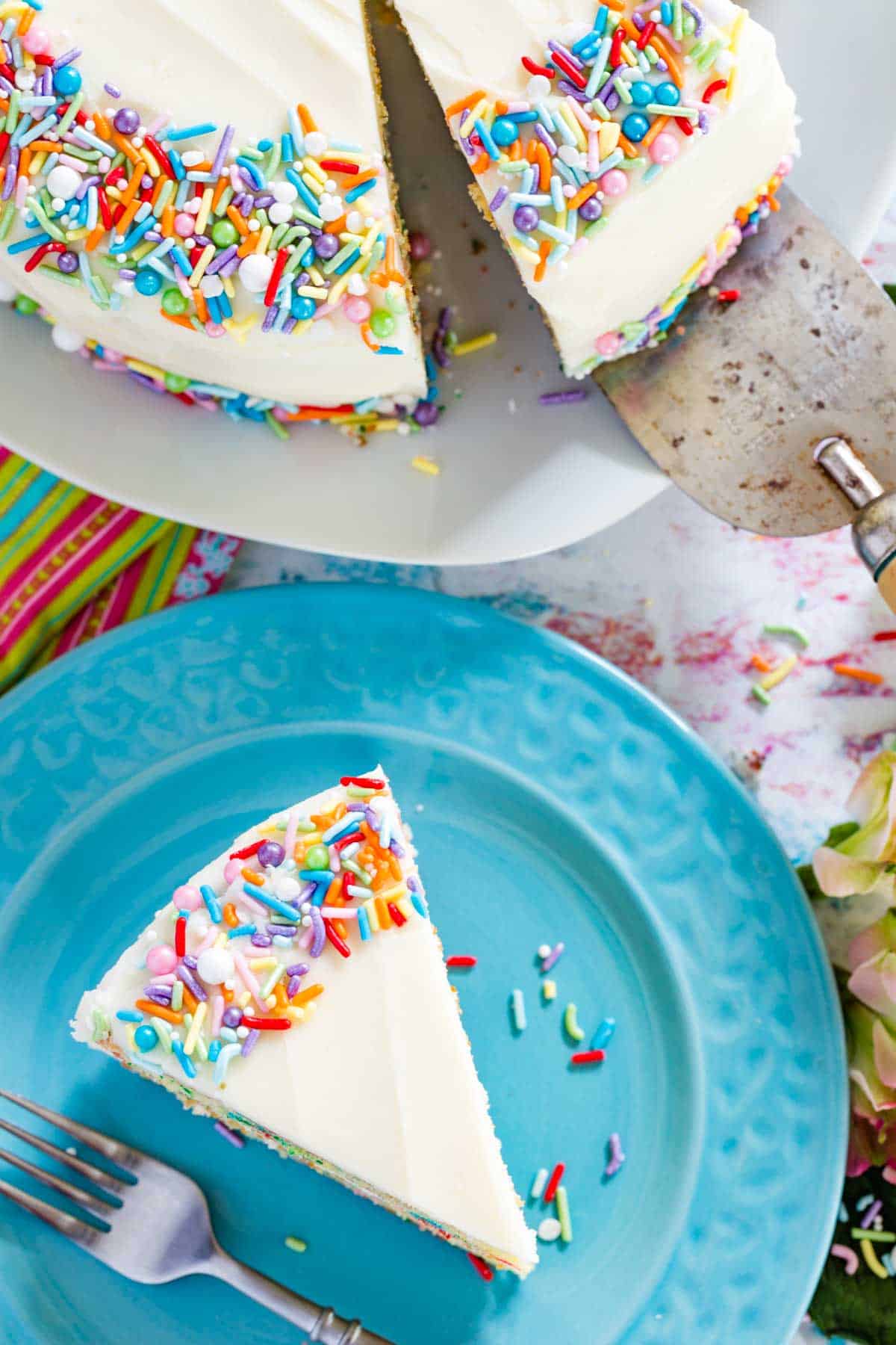 Top view of a slice of gluten free funfetti cake on a blue plate, next to a whole frosted cake.