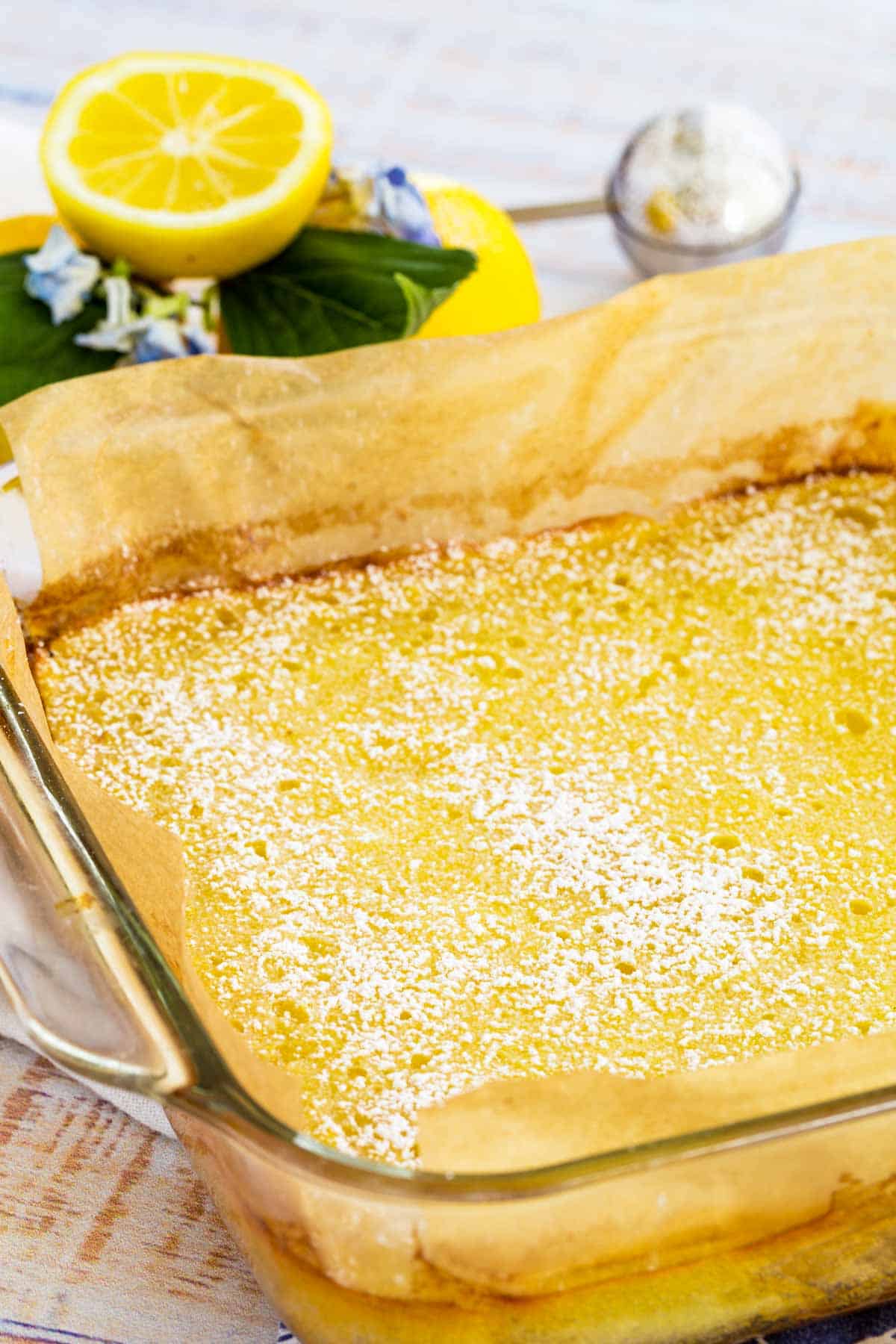 Gluten free lemon bars in a baking dish, dusted with powdered sugar with lemons in the background.