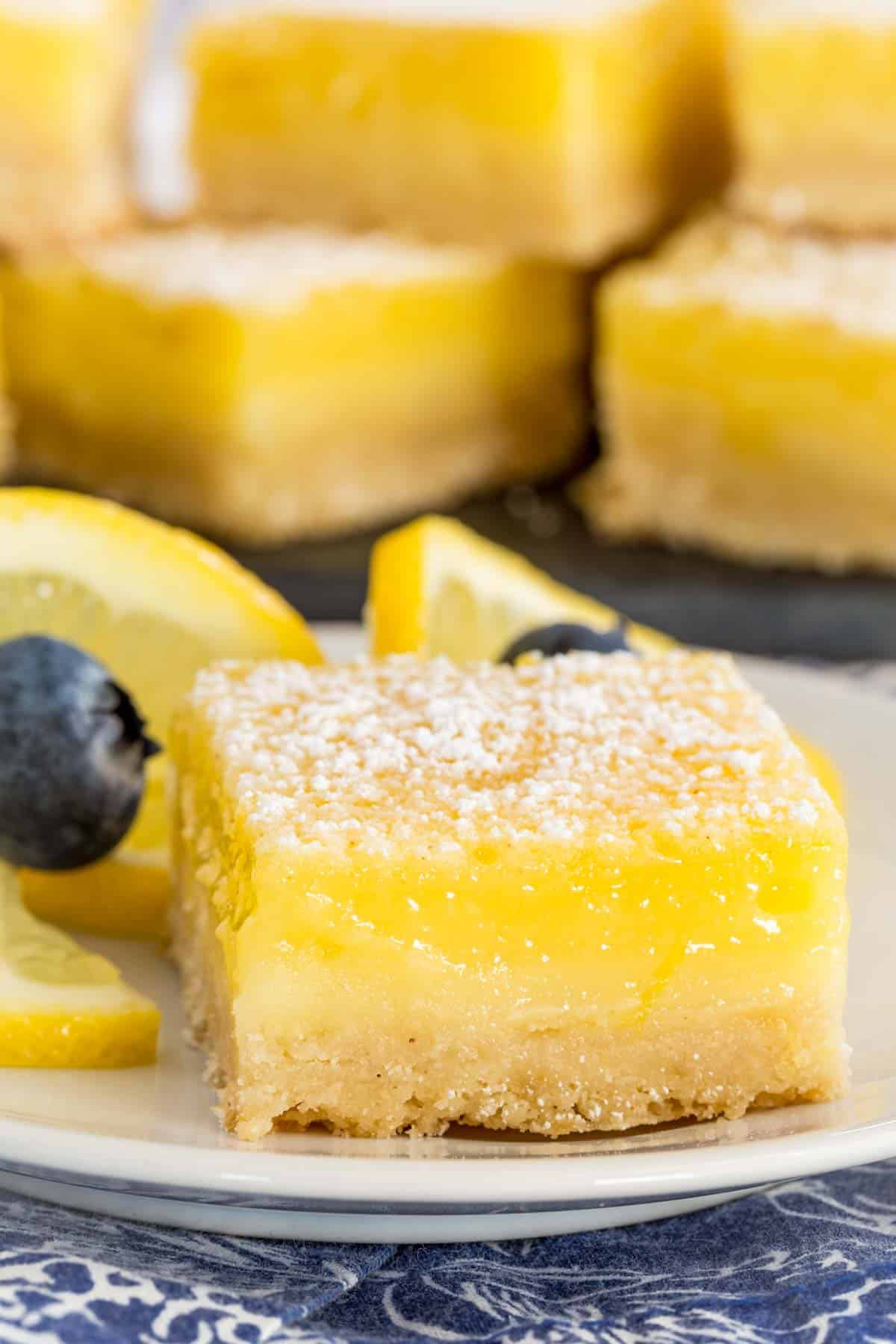 A lemon bar on a plate next to blueberries and lemon slices, with lemon bars in the background.