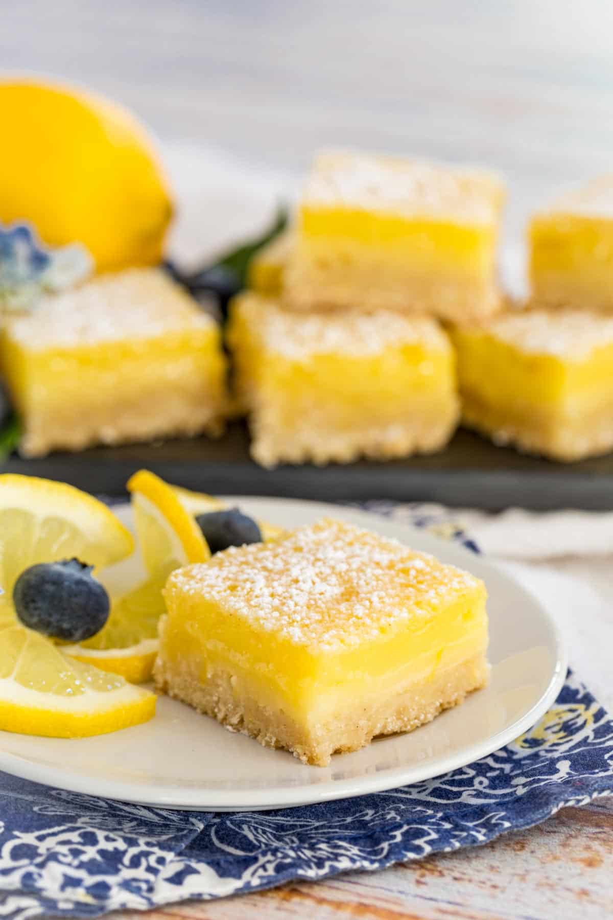 A lemon bar on a plate next to blueberries and lemon slices, with lemon bars in the background.