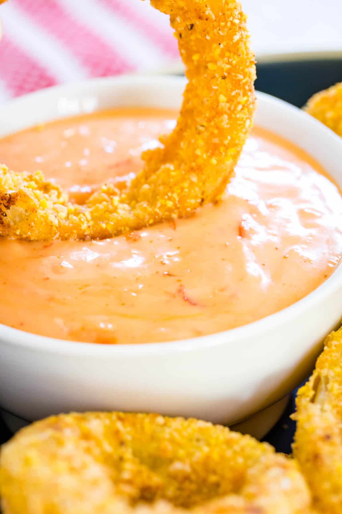 An onion ring being dunked into a small bowl of dipping sauce.