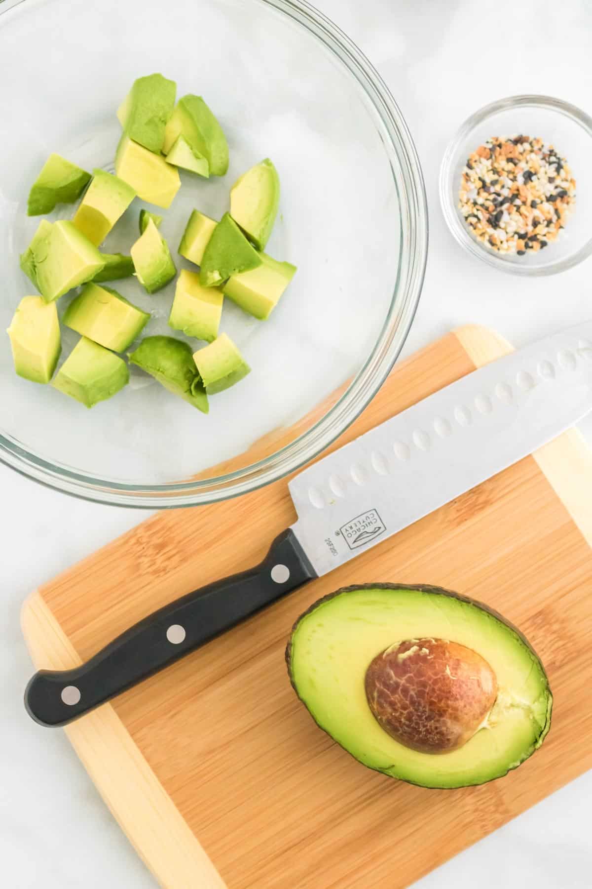 Half an avocado on a wooden cutting board next to a knife, with a bowl of diced avocado near by.