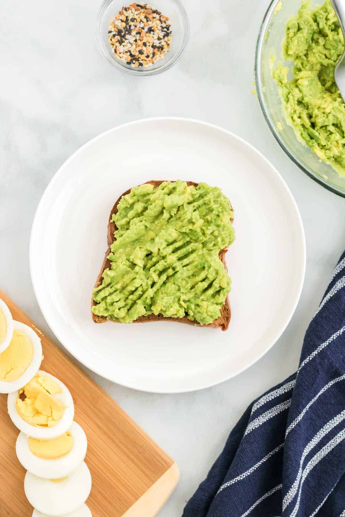 A slice of gluten-free bread topped with mashed avocado on a plate, next to a cutting board with sliced hard boiled eggs.