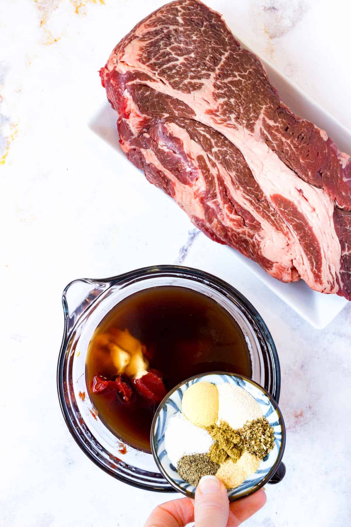 BBQ sauce ingredients are added into a glass mixing bowl next to an uncooked beef chuck roast.