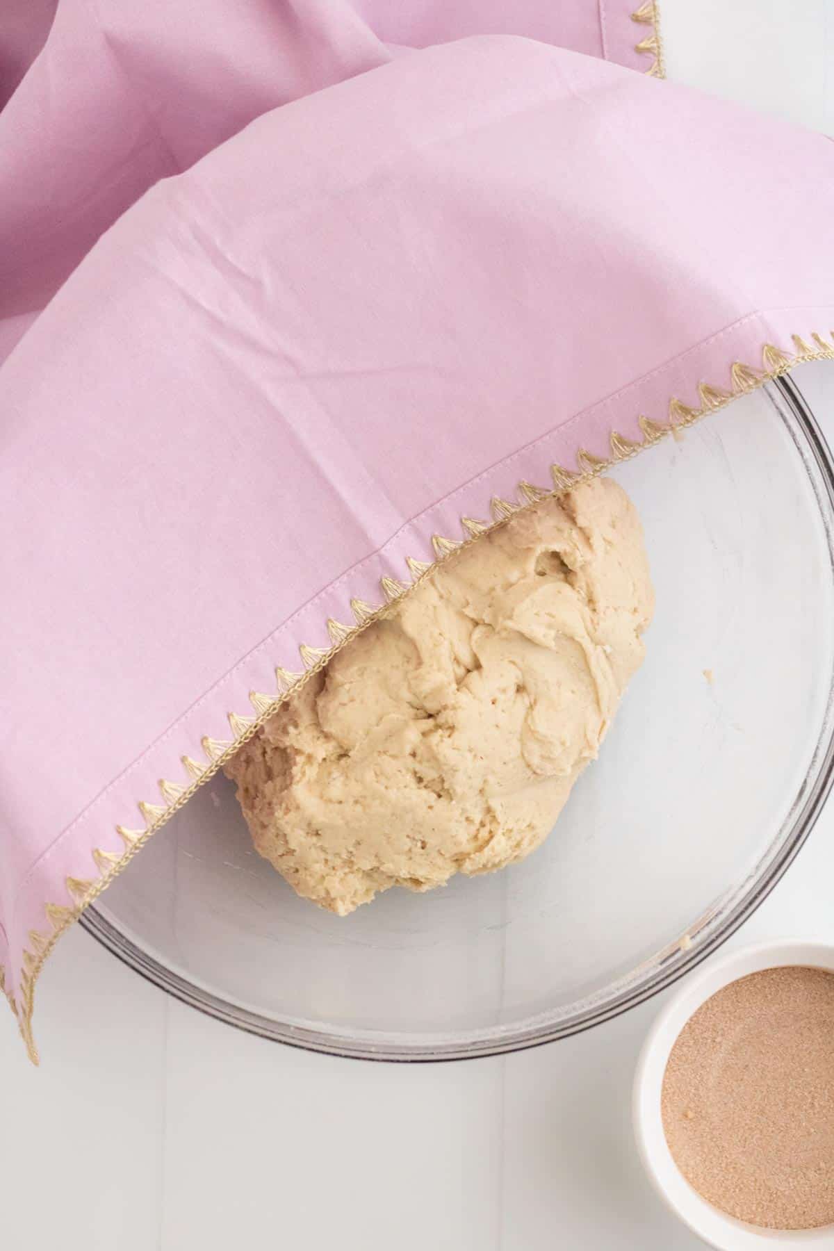 A pink towel partway covering a bowl of dough.