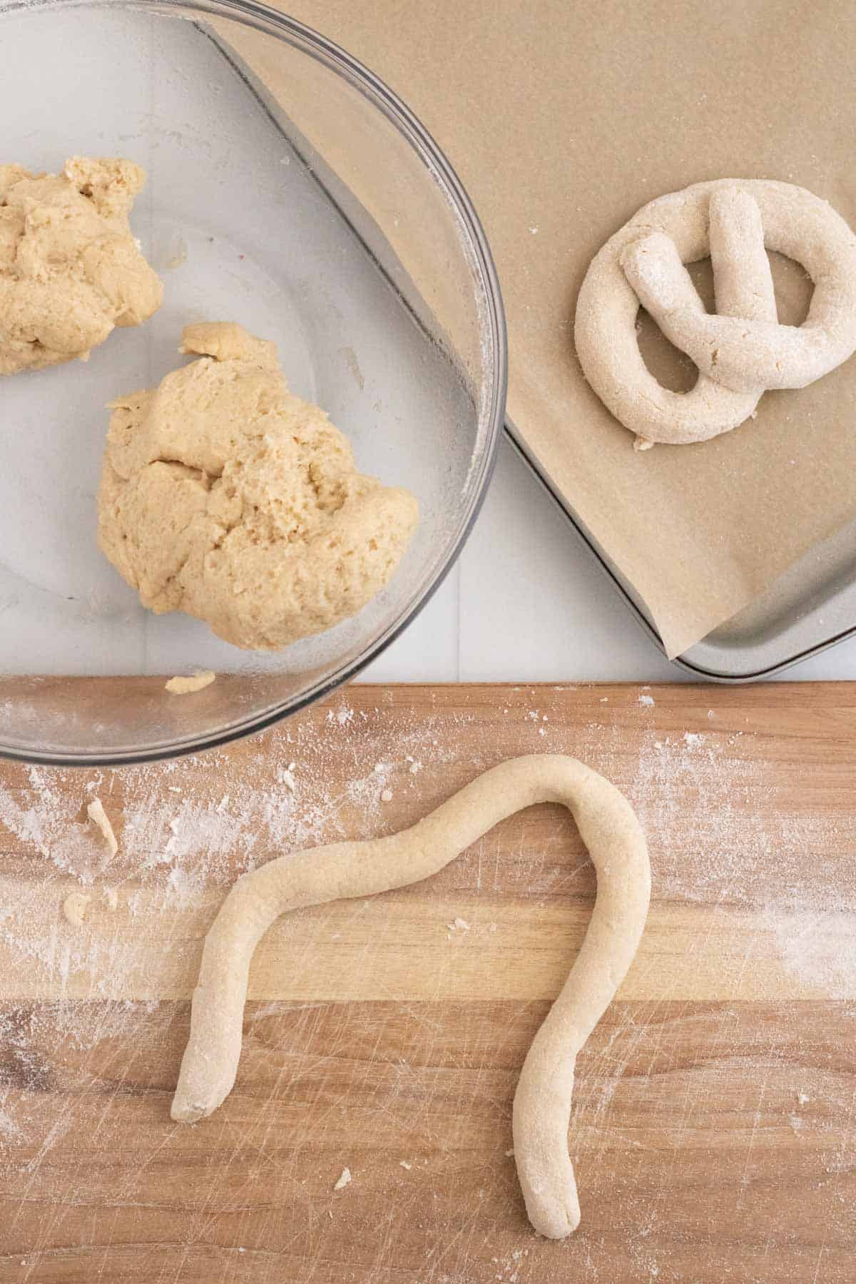 A bowl of pretzel dough, some dough rolled into a rope, and some formed into a pretzel shape.