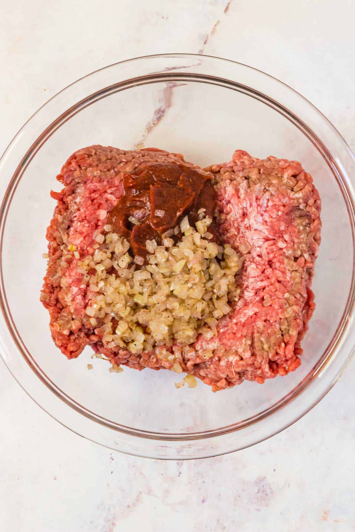 Raw ground beef, ketchup, and sauteed shallots in a glass bowl.