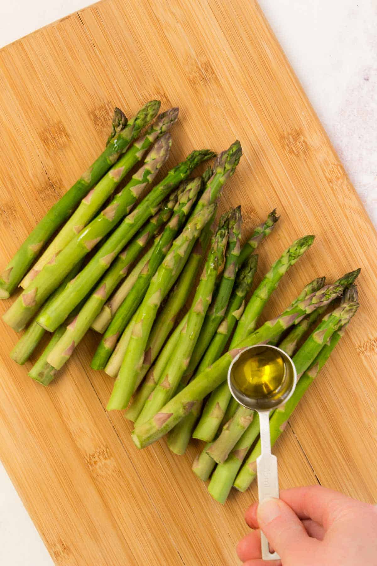 Drizzling olive oil out of a measuring spoon over asparagus spears on a cutting board.