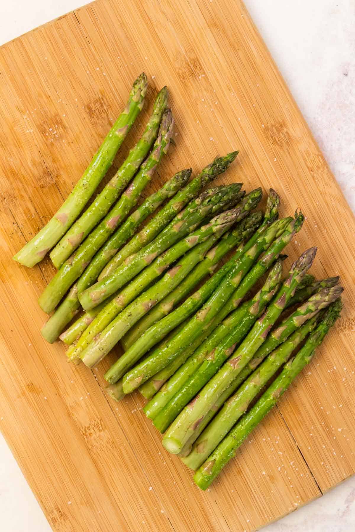 Fresh asparagus spears coated in olive oil, salt, and pepper on a cutting board.