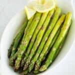 An oval serving dish with lined up oven roasted asparagus spears and a lemon garnish.
