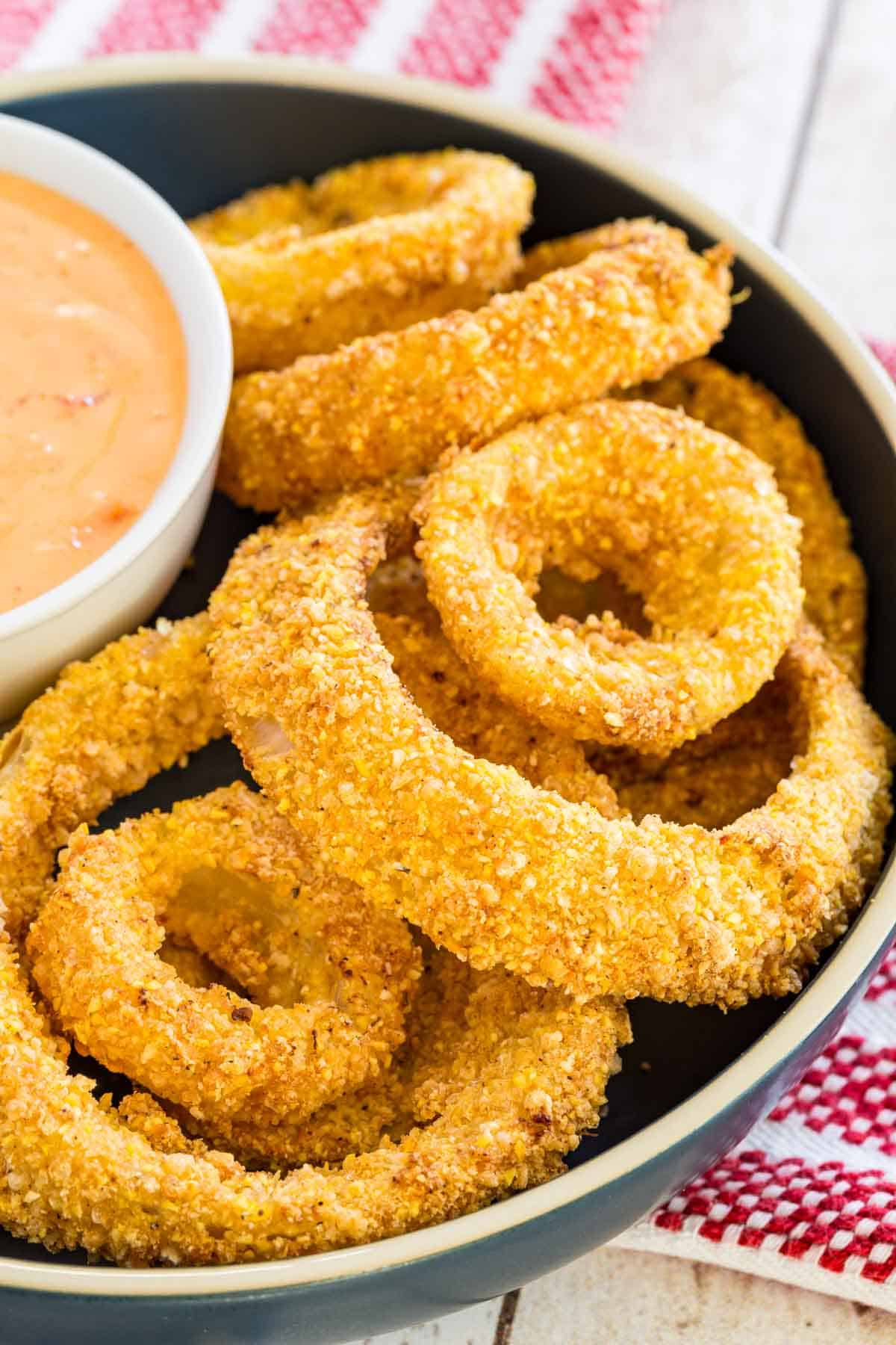 Gluten free air fryer onion rings in a round serving dish around a bowl of dipping sauce.