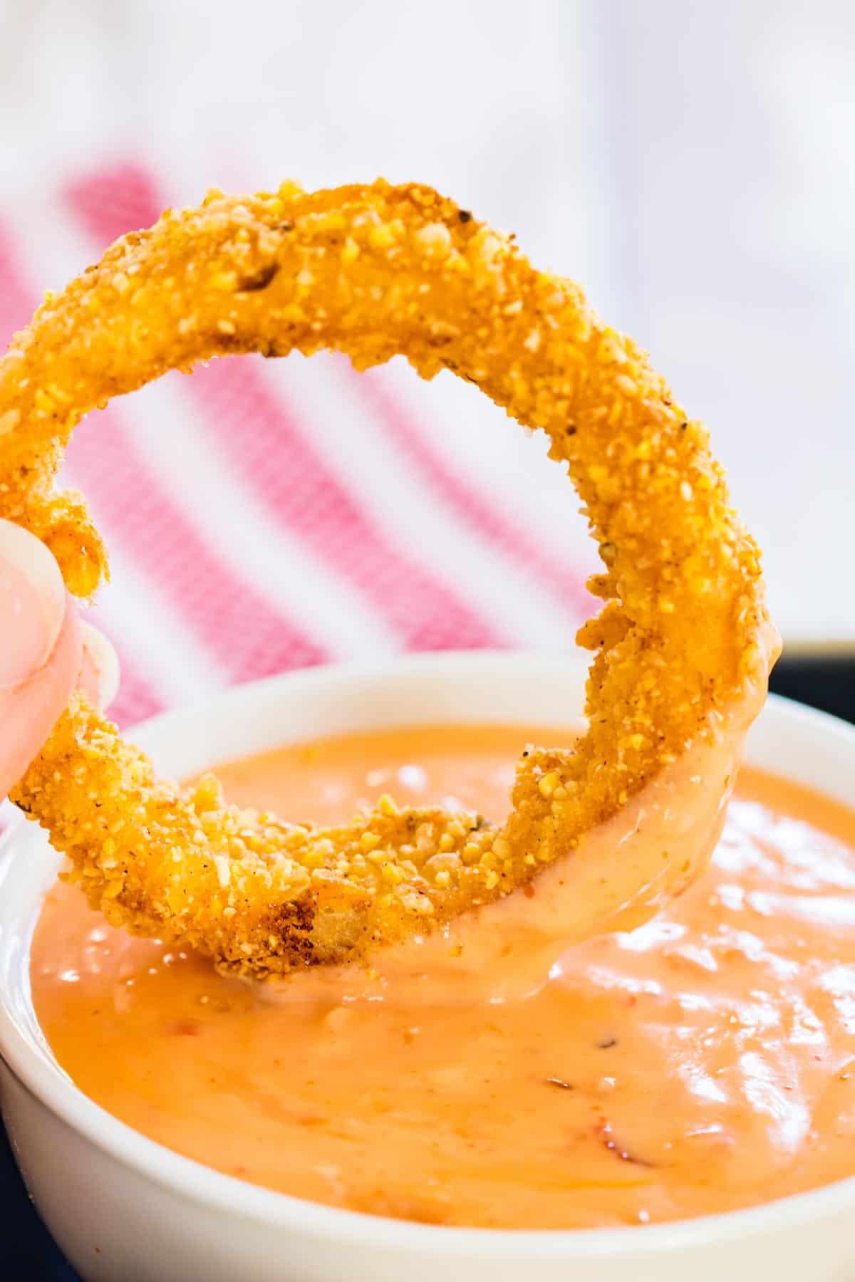 Fingers dipping a crispy air fryer onion ring into a bowl of dipping sauce.
