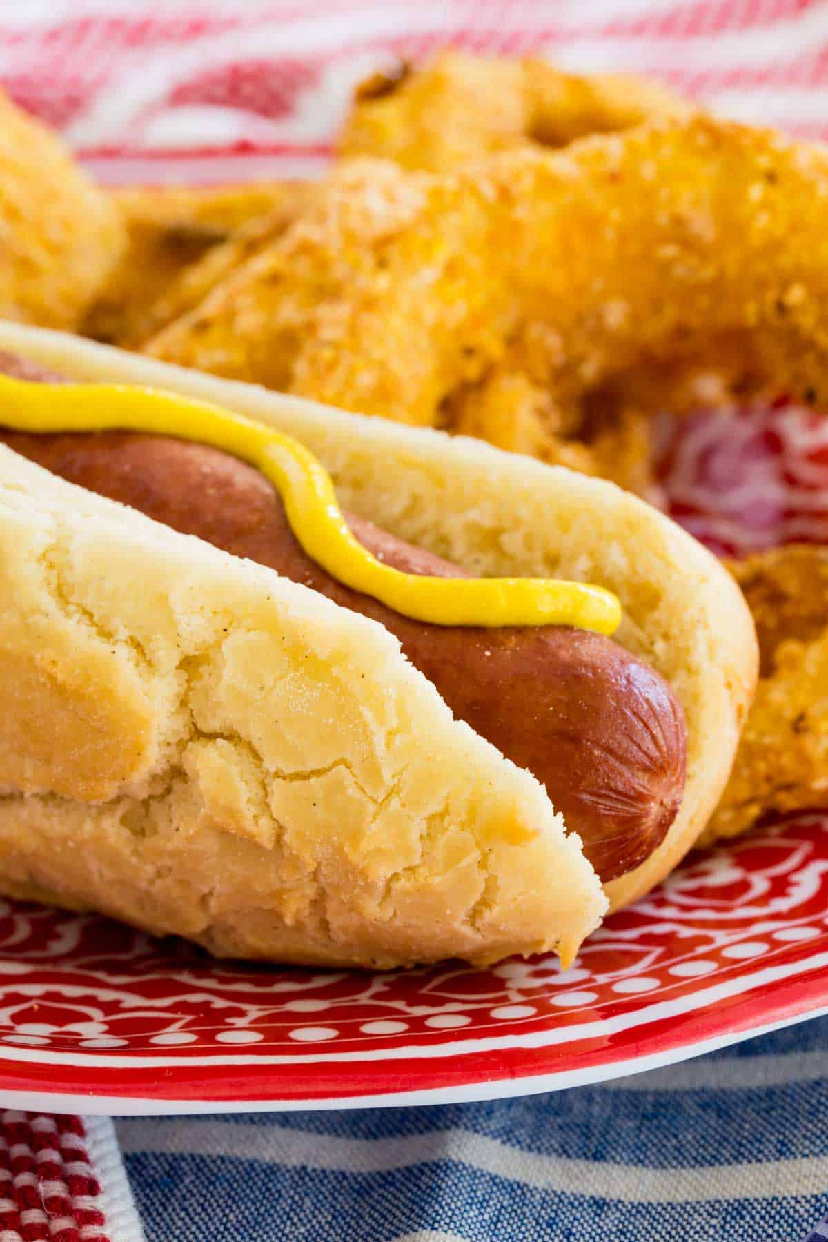 A gluten free hot dog topped with yellow mustard, served on a plate next to onion rings.