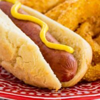 A gluten free hot dog topped with yellow mustard, served on a plate next to onion rings.